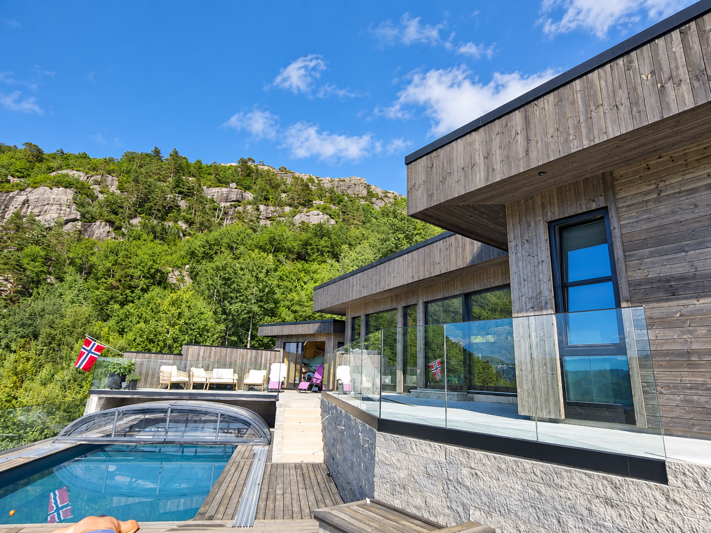 Moderne hus med glassrekkverk, terrasse og basseng, omgitt av skogkledde berg